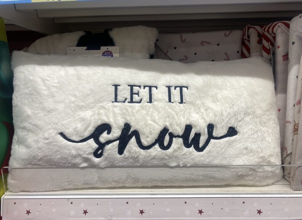 White fluffy cushion with the words 'Let It Snow' embroidered in dark lettering on a shop shelf.